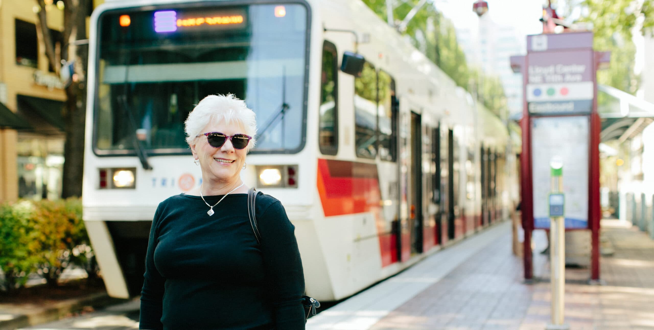 An older woman wearing sunglasses smiles in front of a streetcar.